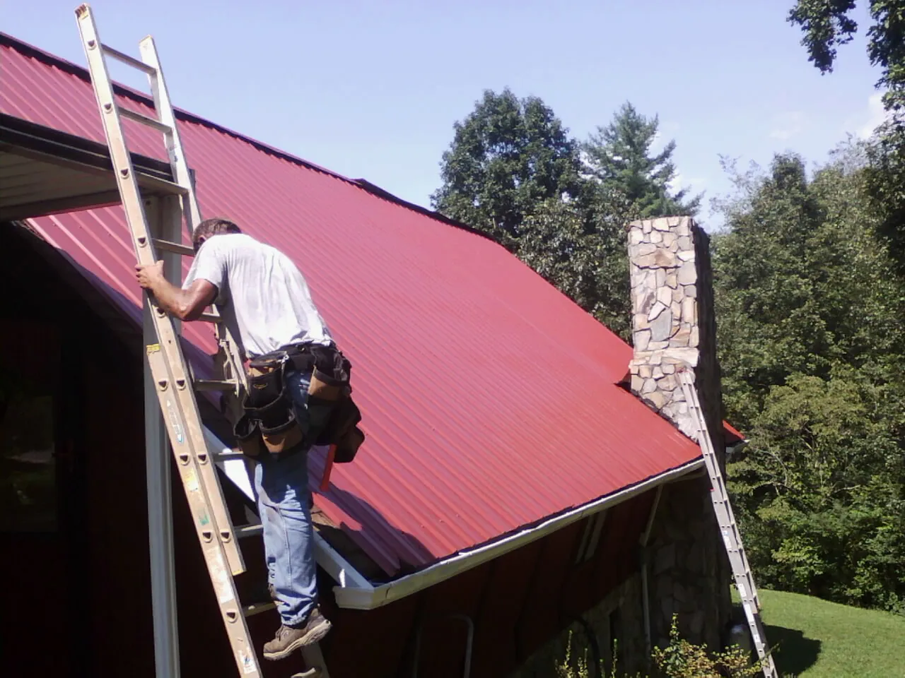 Roofer on a ladder beside a red metal roof and stone chimney — Soffit & Fascia Repair by Roofing Brothers in Broad Brook