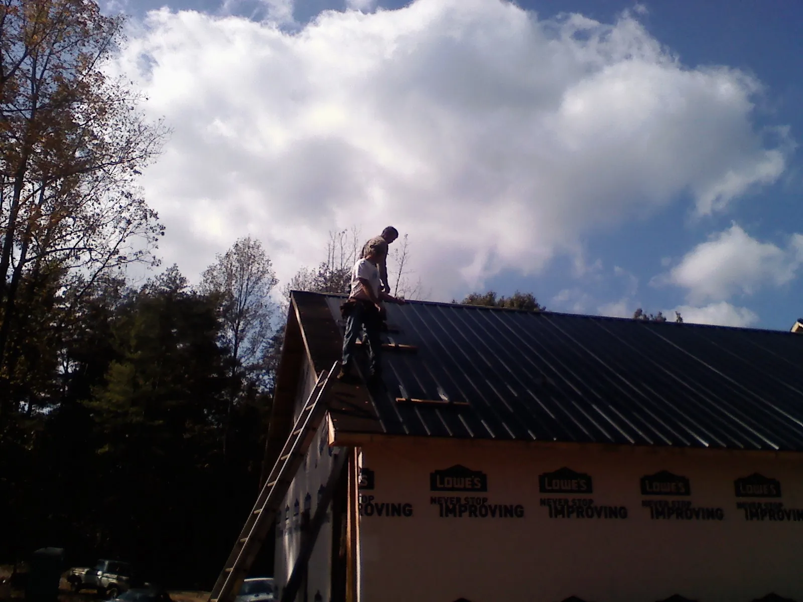 Roofer fastening dark metal panels high against a cloudy sky, from a Roof Inspection job in Broad Brook